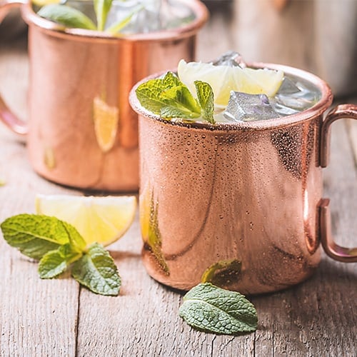 2 unlined copper mugs holding moscow mules surrounded by mint leaves