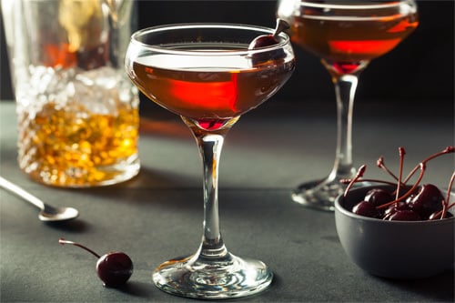 Two Manhattan cocktails next to a bowl of brandied cherries