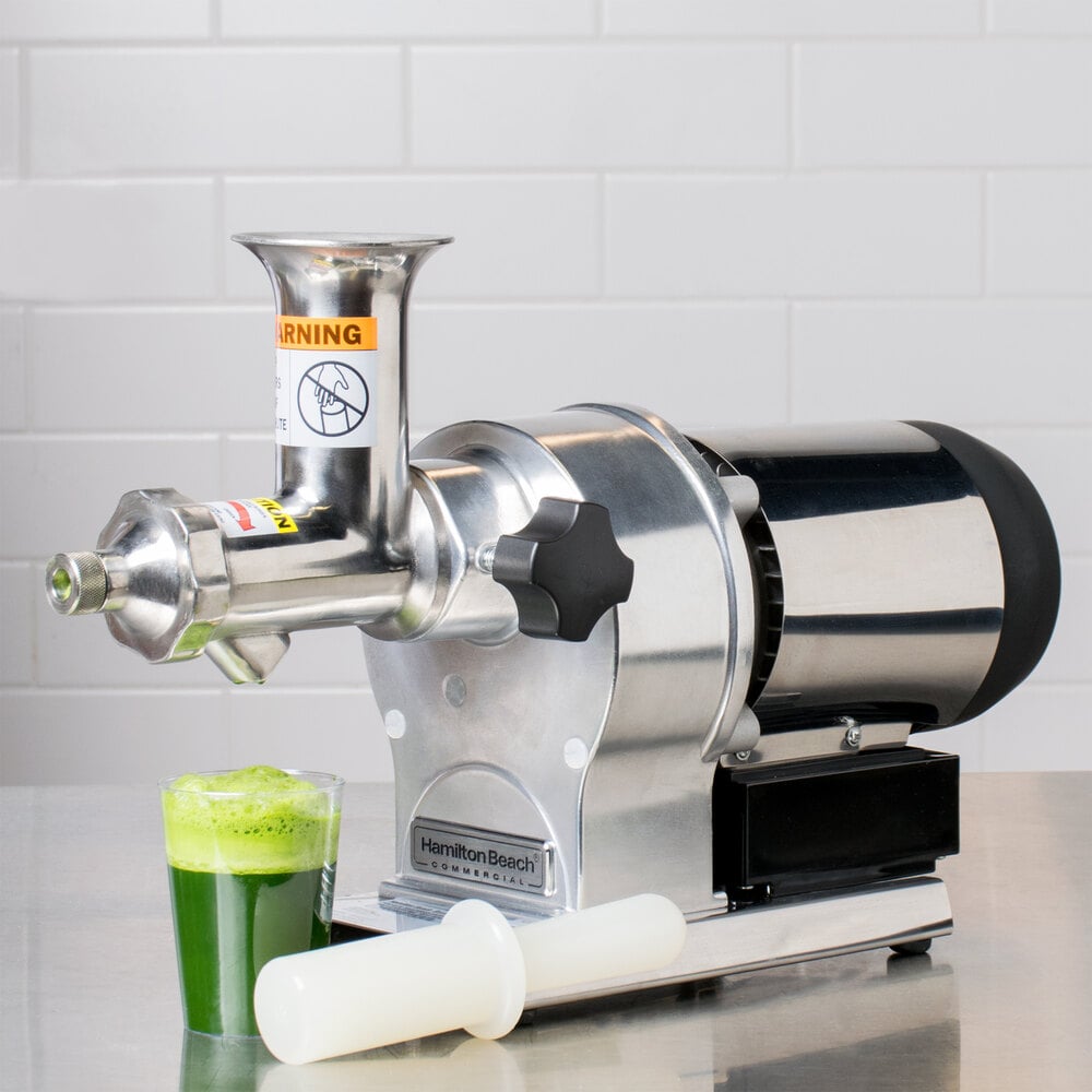 Wheatgrass juicer sitting on a stainless steel table with a glass of green juice and a white pusher