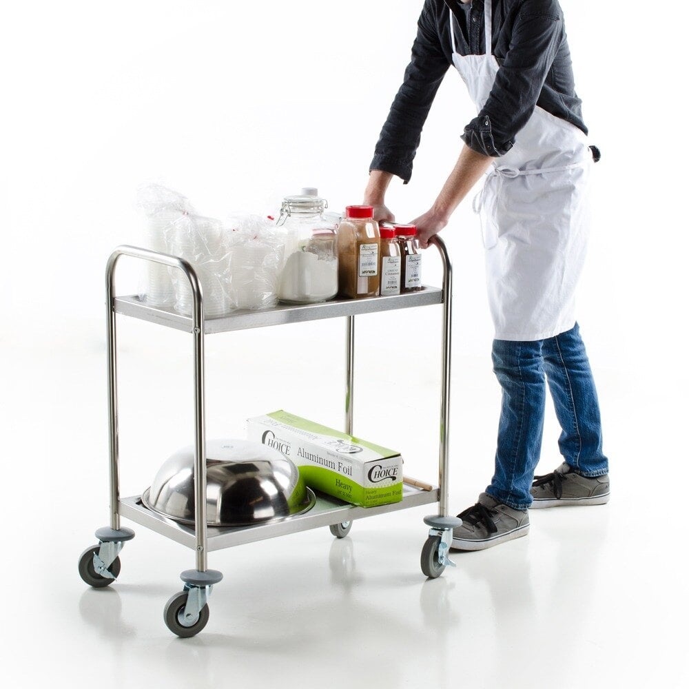 Man with white apron pushing a two tier metal utility cart with various foodservice items