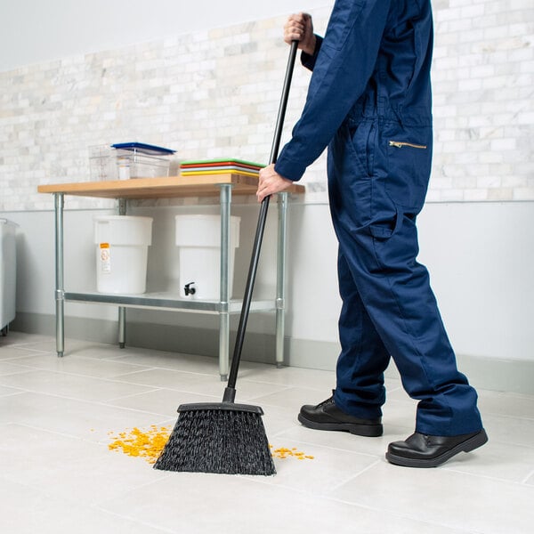 A warehouse broom with black unflagged bristles and a 48-inch handle being used to sweep debris on a tiled floor.