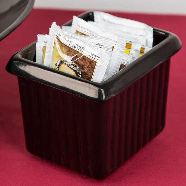 A Tablecraft black rectangular server with ridges holding packets of sugar on a hotel buffet counter.