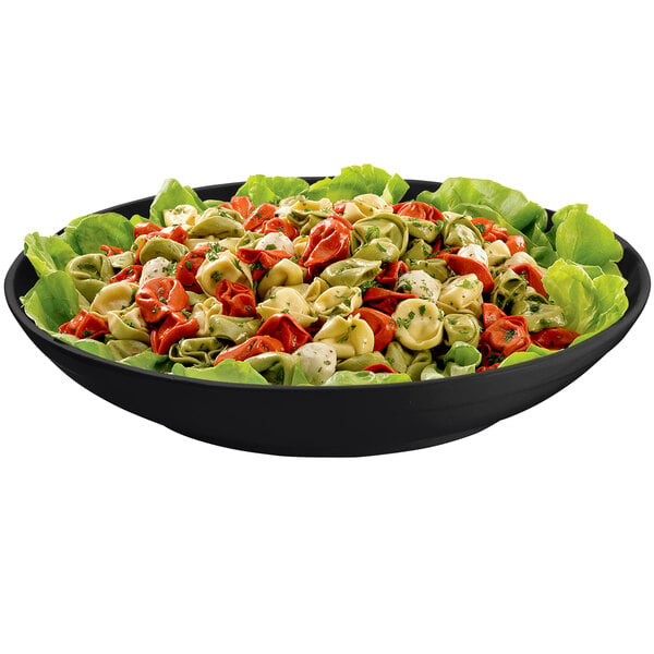 A Tablecraft black cast aluminum pasta bowl filled with pasta and lettuce on a table with lettuce and tortellini.