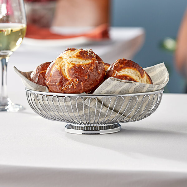 A Tablecraft chrome oval basket with bread rolls inside on a table.