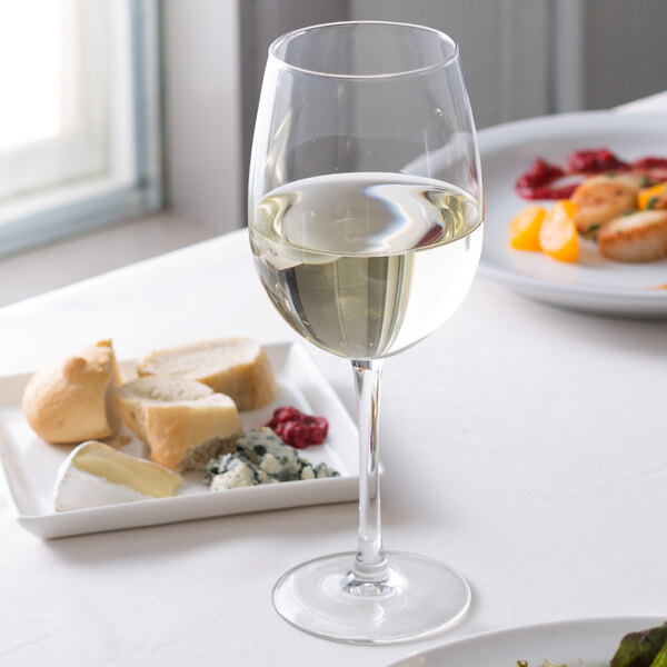 An Arcoroc tall wine glass filled with white wine next to a plate of food on a table.