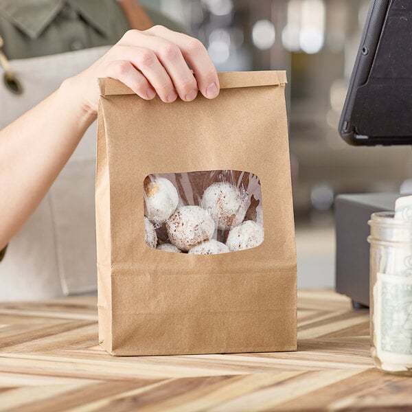 A person holding a brown Kraft paper bag with a clear window filled with cookies.