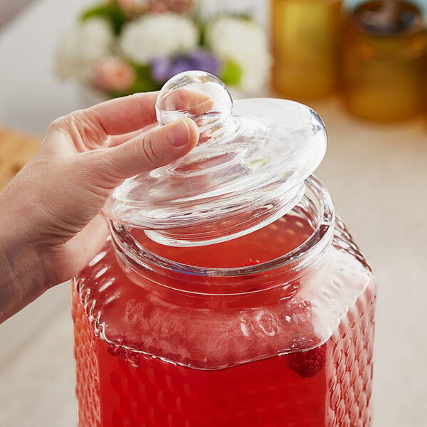 A hand pouring red liquid into an Acopa glass beverage dispenser on a table.