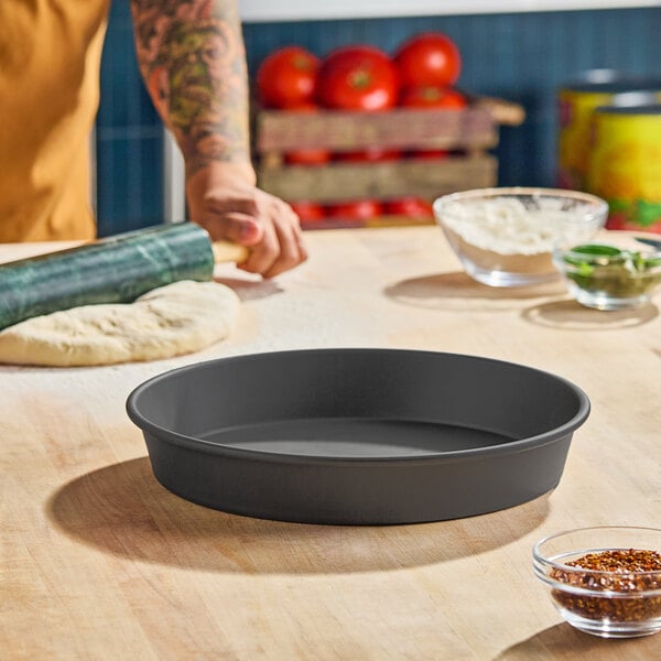 A round, hard coat anodized aluminum pizza pan with tapered sides, measuring 12 inches in diameter and 2 inches deep.
