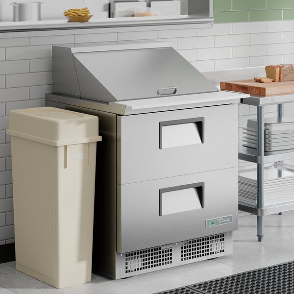 A stainless steel two-drawer mega top refrigerated sandwich prep table with a closed lid, positioned next to a beige trash bin in a commercial kitchen setting.