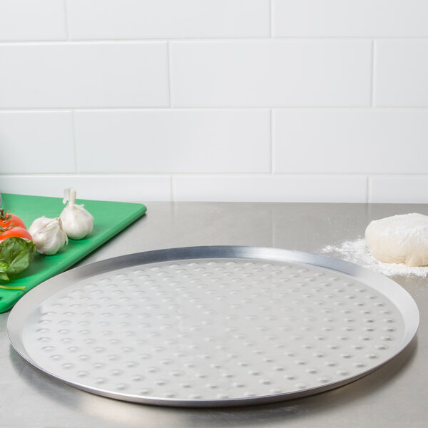 An American Metalcraft pizza pan with pizza dough and ingredients on a counter.