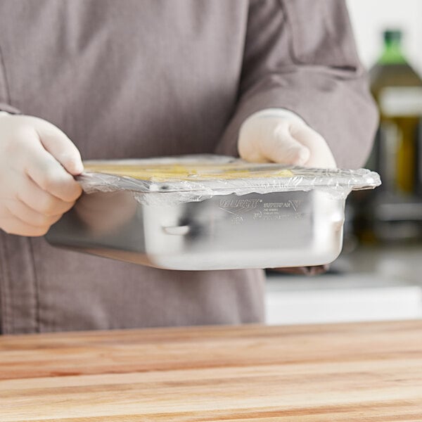 A person in a chef's uniform holding a Vollrath stainless steel pan of food.