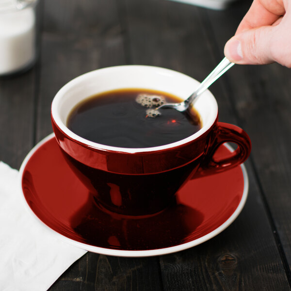 A hand holding a spoon in a red CAC Venice cup of coffee.