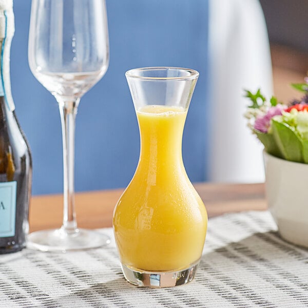 A glass jar filled with yellow liquid sits on a table with wine glasses and a glass of orange juice.