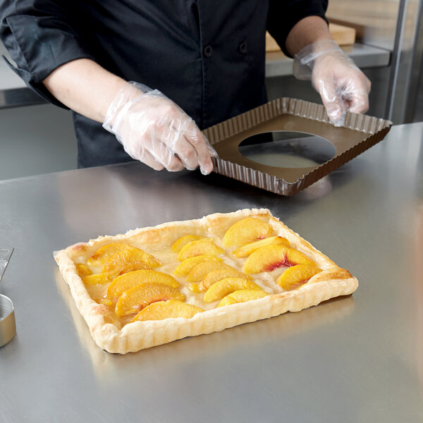 A person in plastic gloves prepares a peach tart in a Gobel fluted rectangle quiche pan.