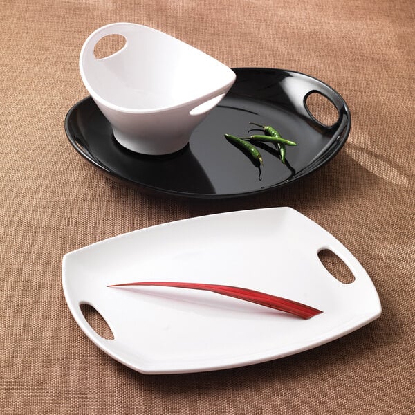 An Elite Global Solutions black melamine oval platter with handles holding a white bowl and black plate with a red pepper on it.