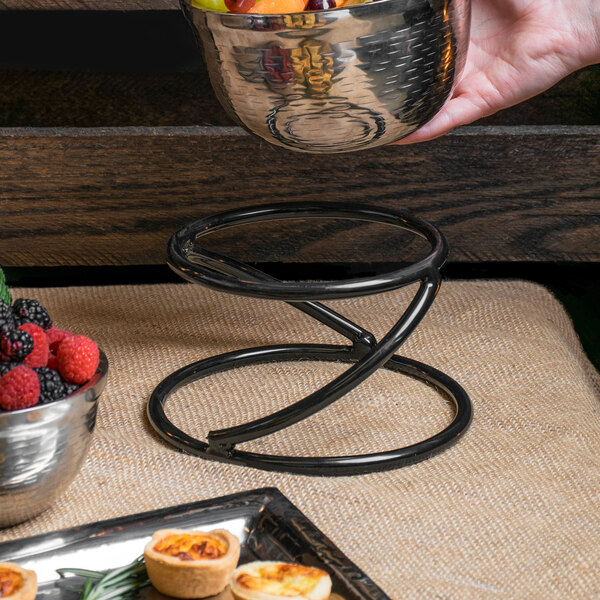 A hand uses an Elite Global Solutions round metal stand to hold a bowl of fruit on a table.