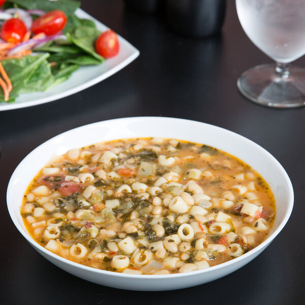 A 10 Strawberry Street white porcelain soup bowl filled with soup, pasta, and vegetables on a table with a glass of water.