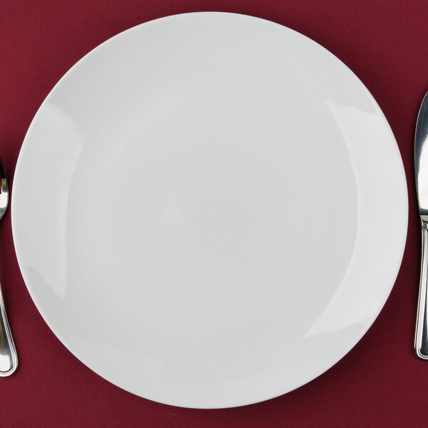 A white 10 Strawberry Street porcelain charger plate with a knife and fork on a red surface.