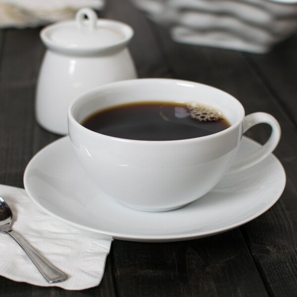 A white porcelain cup of coffee and saucer on a table with a spoon.