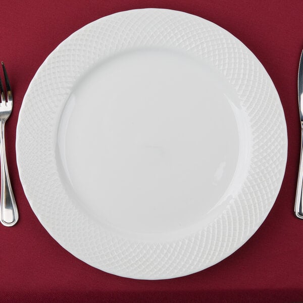 A white 10 Strawberry Street Wicker porcelain charger plate with a fork and knife on a red tablecloth.