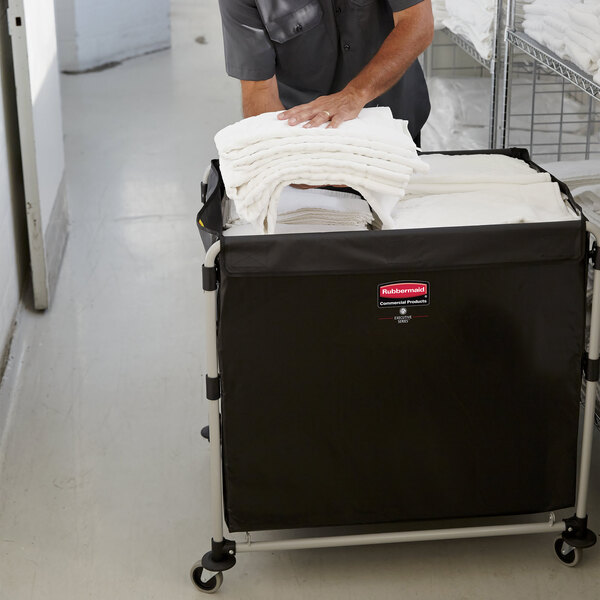 A man wearing a black shirt pushing a black Rubbermaid laundry cart full of white towels.