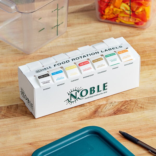 A white Noble Products dispenser with 7 different colored date labels on a wooden surface.