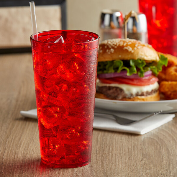 A red customizable plastic tumbler filled with red liquid and ice with a straw next to a plate of food including a burger