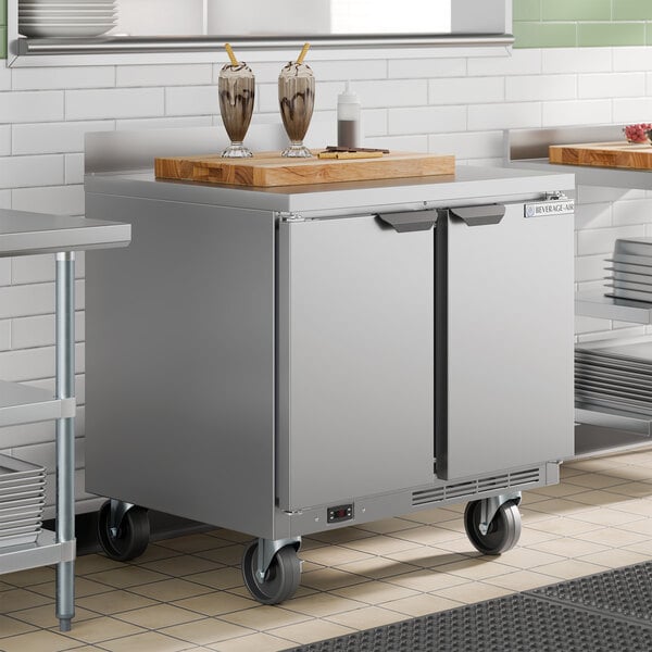 A stainless steel two-door worktop freezer on wheels with a cutting board and milkshakes on top.