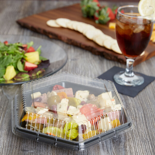 A Genpak clear dome lid on a plastic container of salad on a table next to a glass of drink.