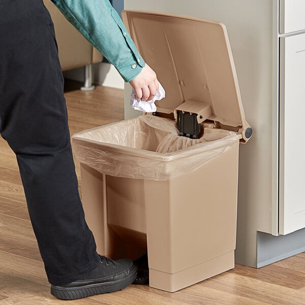 A person throwing paper into a Rubbermaid plastic trash can.