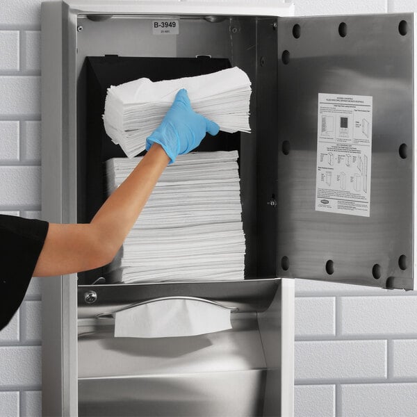 A hand in a blue glove putting paper towels into a metal box with a sign for a Bobrick paper towel dispenser.
