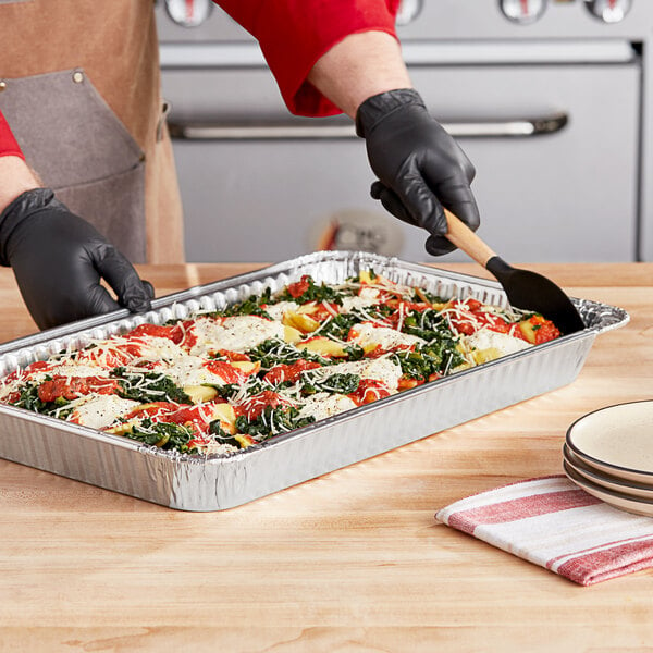 A person in black gloves using a spatula to serve food from a Western Plastics foil steam table pan.