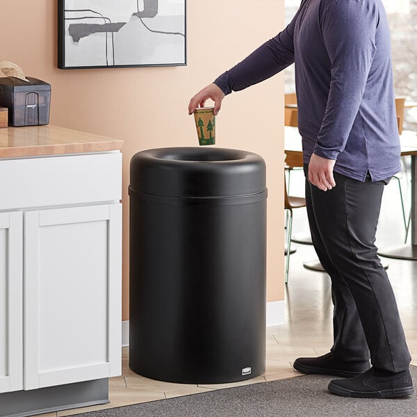 A man in a purple shirt throwing a cup into a Rubbermaid Crowne round open top trash can.