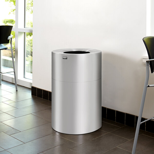 A silver Rubbermaid Atrium waste receptacle in a room.
