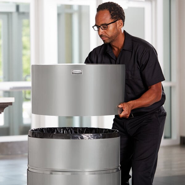 A man in a black shirt and glasses holding a large silver Rubbermaid waste receptacle.
