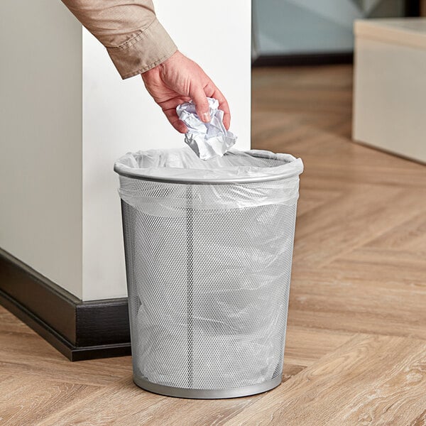 A person throws crumpled paper into a Rubbermaid mesh wastebasket.