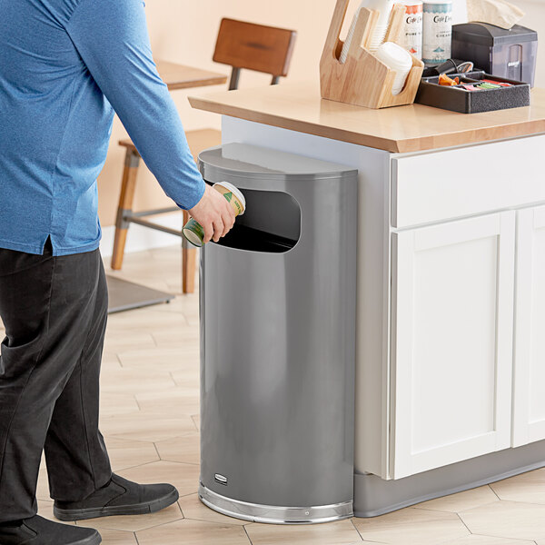A man standing next to a Rubbermaid half round trash can holding a coffee cup.