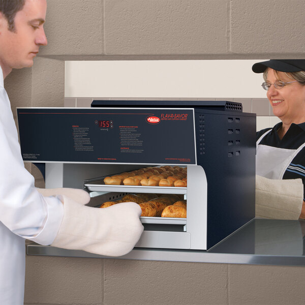 A man and woman wearing aprons use a Hatco Flav-R-Savor Pass-Through Heated Air Curtain to put bread in an oven.