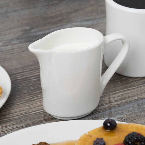 A white Tuxton China creamer with a handle.