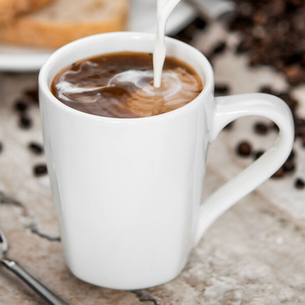 A close-up of a Tuxton white coffee mug with brown liquid and milk being poured into it.