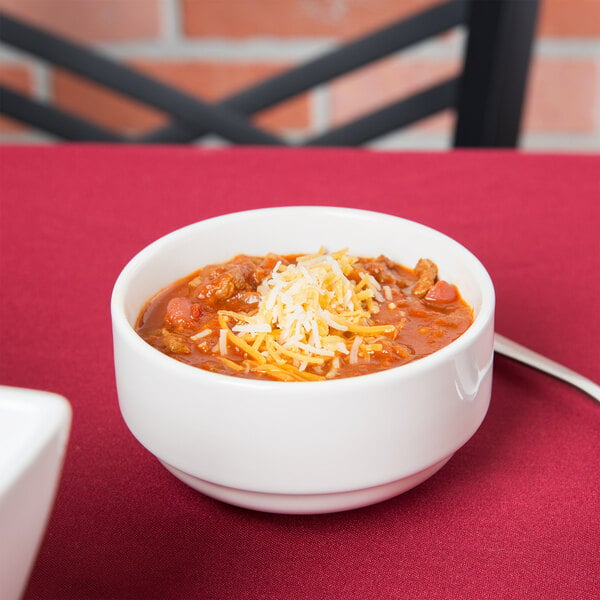 A bowl of chili with cheese and meat in a Tuxton white soup cup on a table.