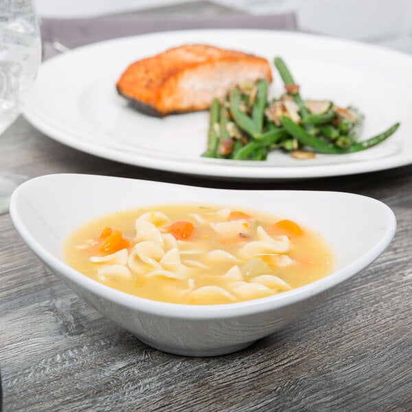 A Tuxton bright white china bowl filled with soup with noodles and carrots on a table with a blurry plate of food.