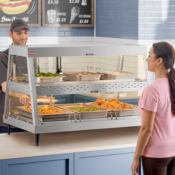 A woman in a pink shirt standing in front of a Hatco stainless steel countertop hot food merchandiser.