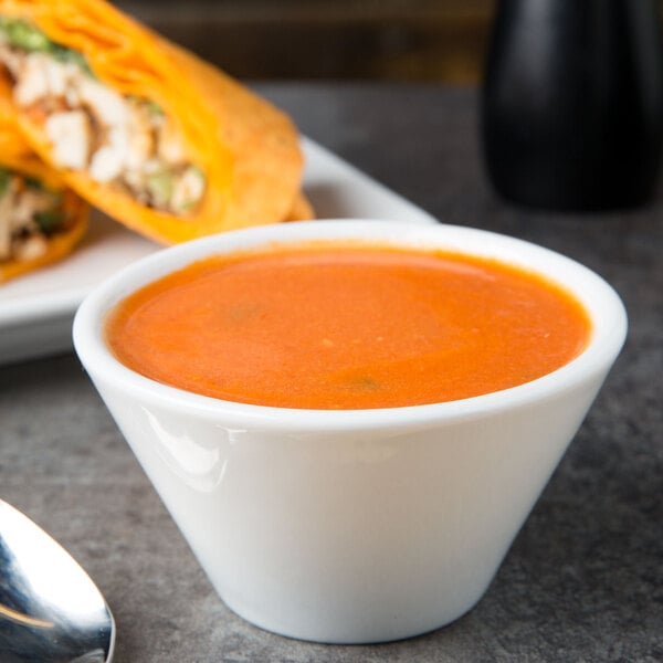 A close up of a TuxTrendz bright white China bouillon cup filled with soup next to a spoon.