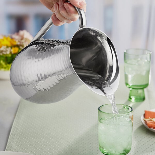 A hand pouring water from a glass into a silver American Metalcraft pitcher.