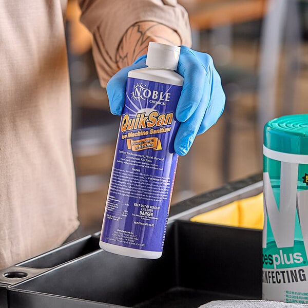 A person in blue gloves holding a bottle of Noble Chemical QuikSan Nickel-Safe ice machine sanitizer.