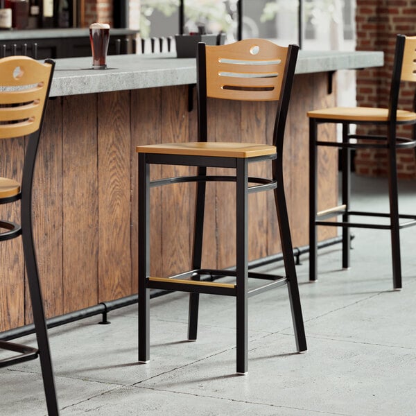 A row of Lancaster Table & Seating wooden bar stools with black frames and natural wood seats and backs sit in front of a bar.