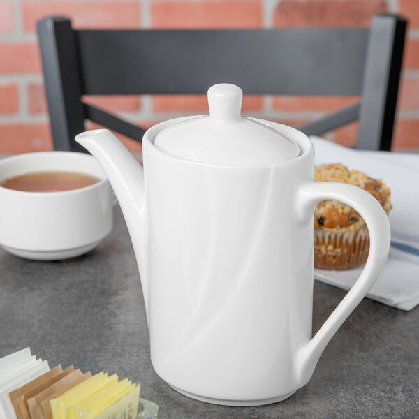 A white Tuxton China coffee pot on a table with a cup of tea.