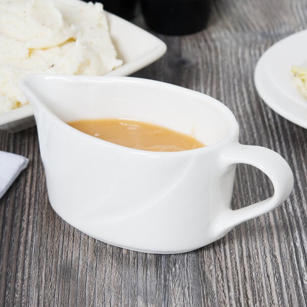 A white Tuxton gravy boat filled with brown liquid on a table with a bowl of mashed potatoes.