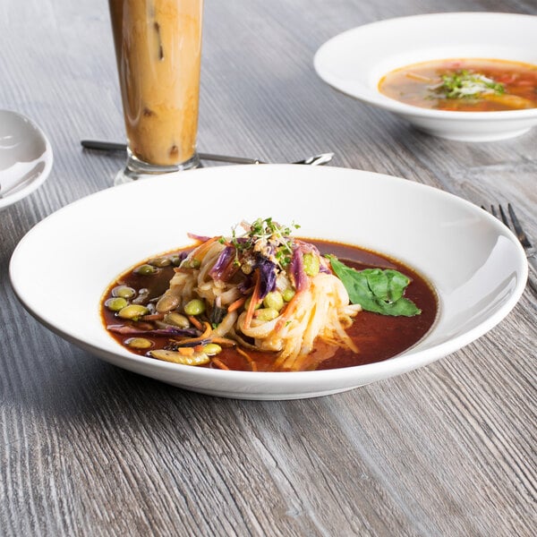 A Tuxton white bowl filled with soup with noodles and vegetables on a table.